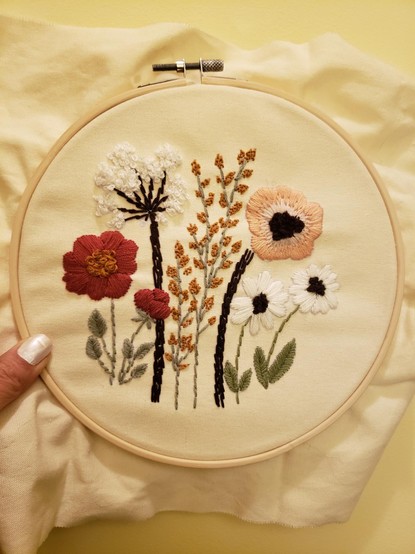 A bunch of flowers and leaves using different types of stitches, in red, pink,  green, brown, white, and gold on a cream background. They are framed by a circular embroidery hoop held by a thumb with white nail polish.